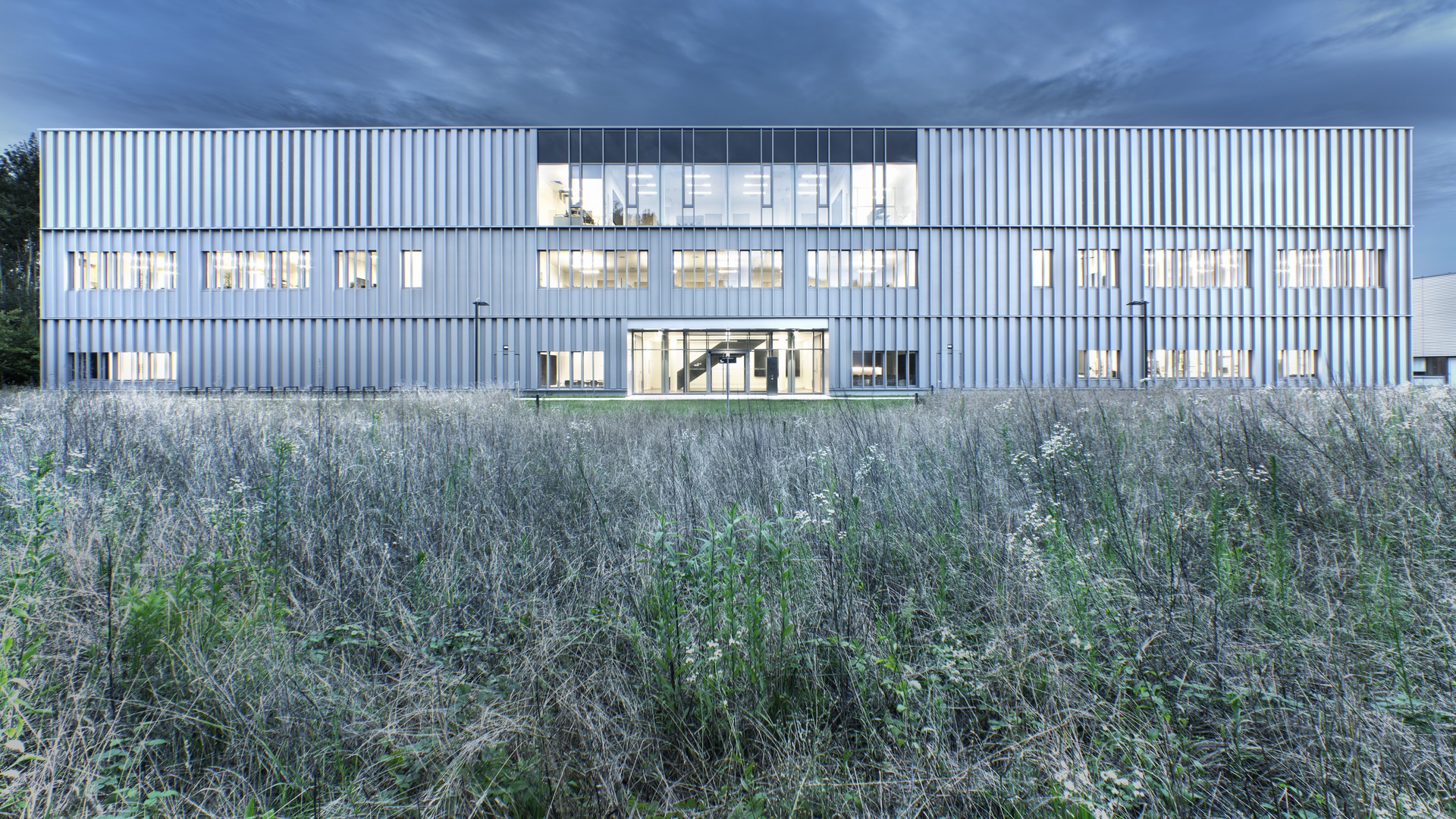 Hightech-Anlage mit großflächiger Verglasung und Metallverkleidung, eingebettet in eine nachhaltige Landschaft unter einem dynamischen, bewölkten Himmel.