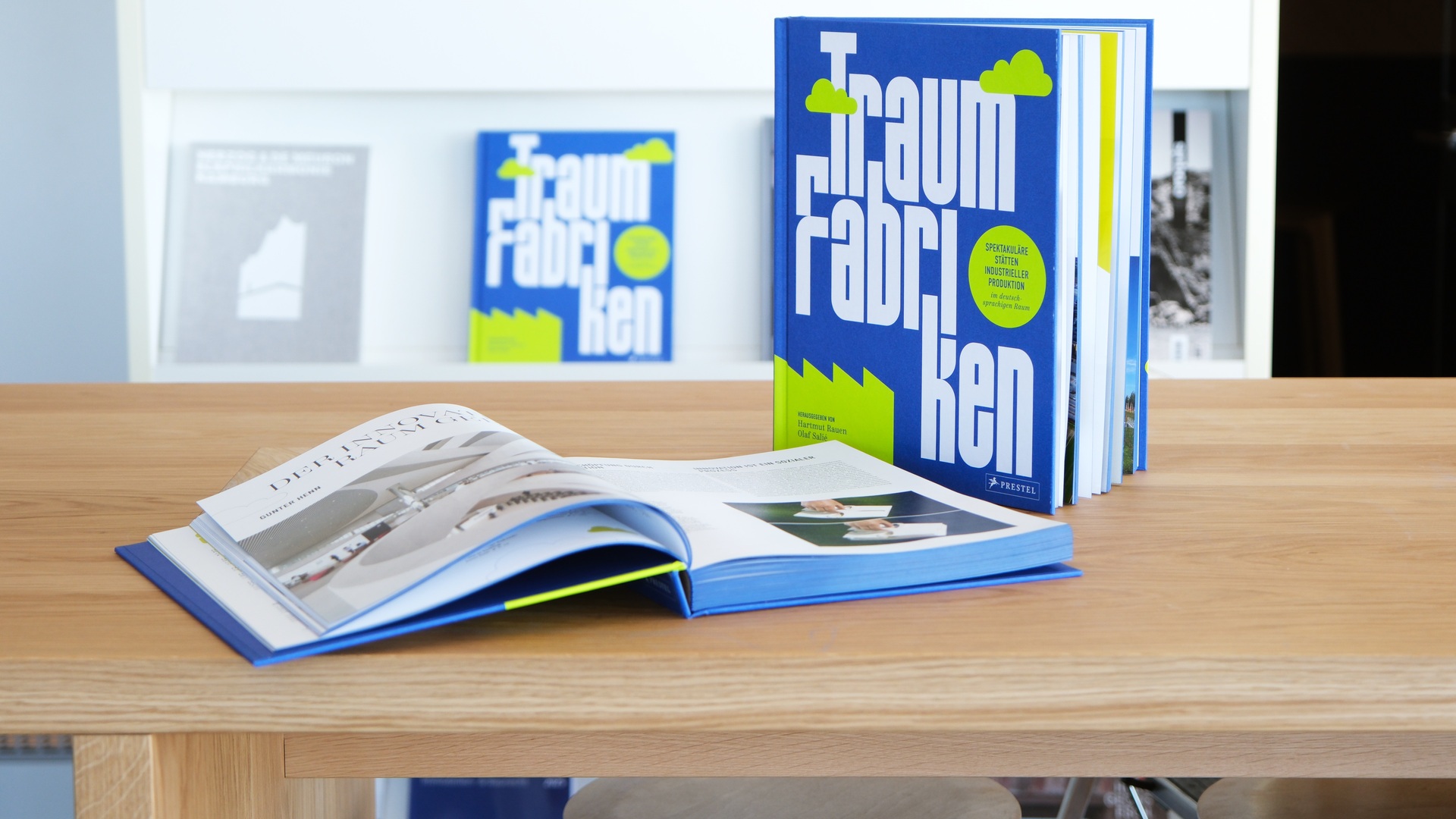 A blue book titled Traum Fabriken stands upright on a table beside an open book, reflecting innovative and functional design inspiration.