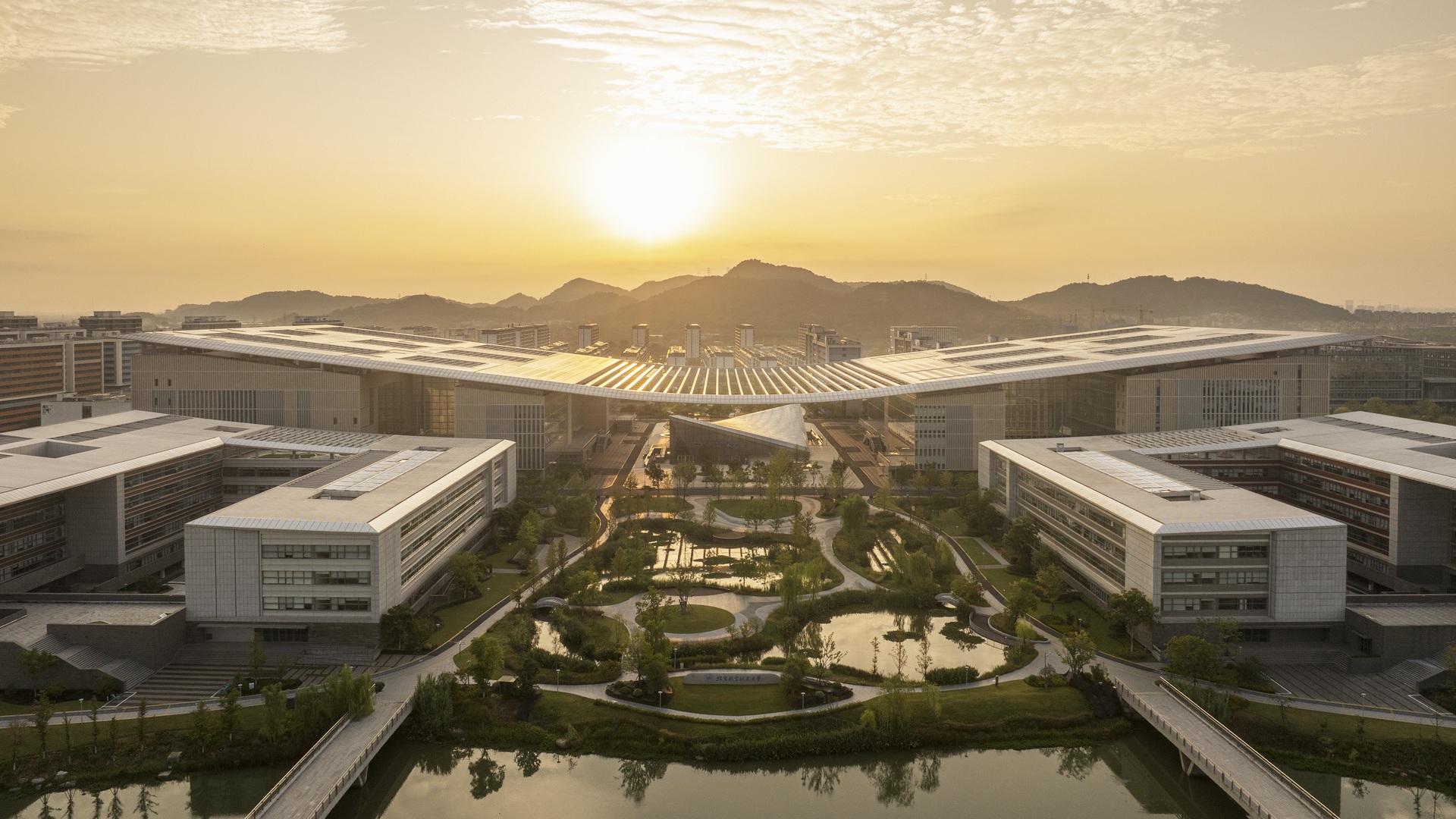 Ein Blick bei Sonnenaufgang auf einen Hightech-Campus mit symmetrischen, umweltfreundlichen Gebäuden, angelegten Gärten und einem spiegelnden Teich.