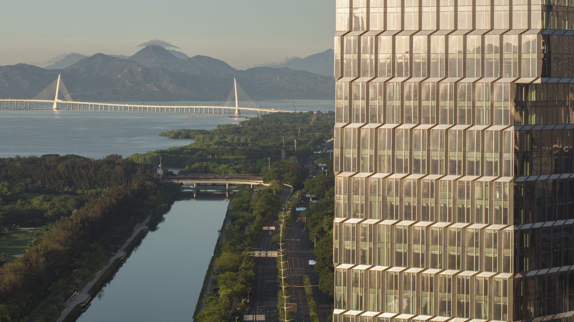 Innovative glass tower by the river, showcasing sustainable, high-tech architecture against a backdrop of bridge and mountains.