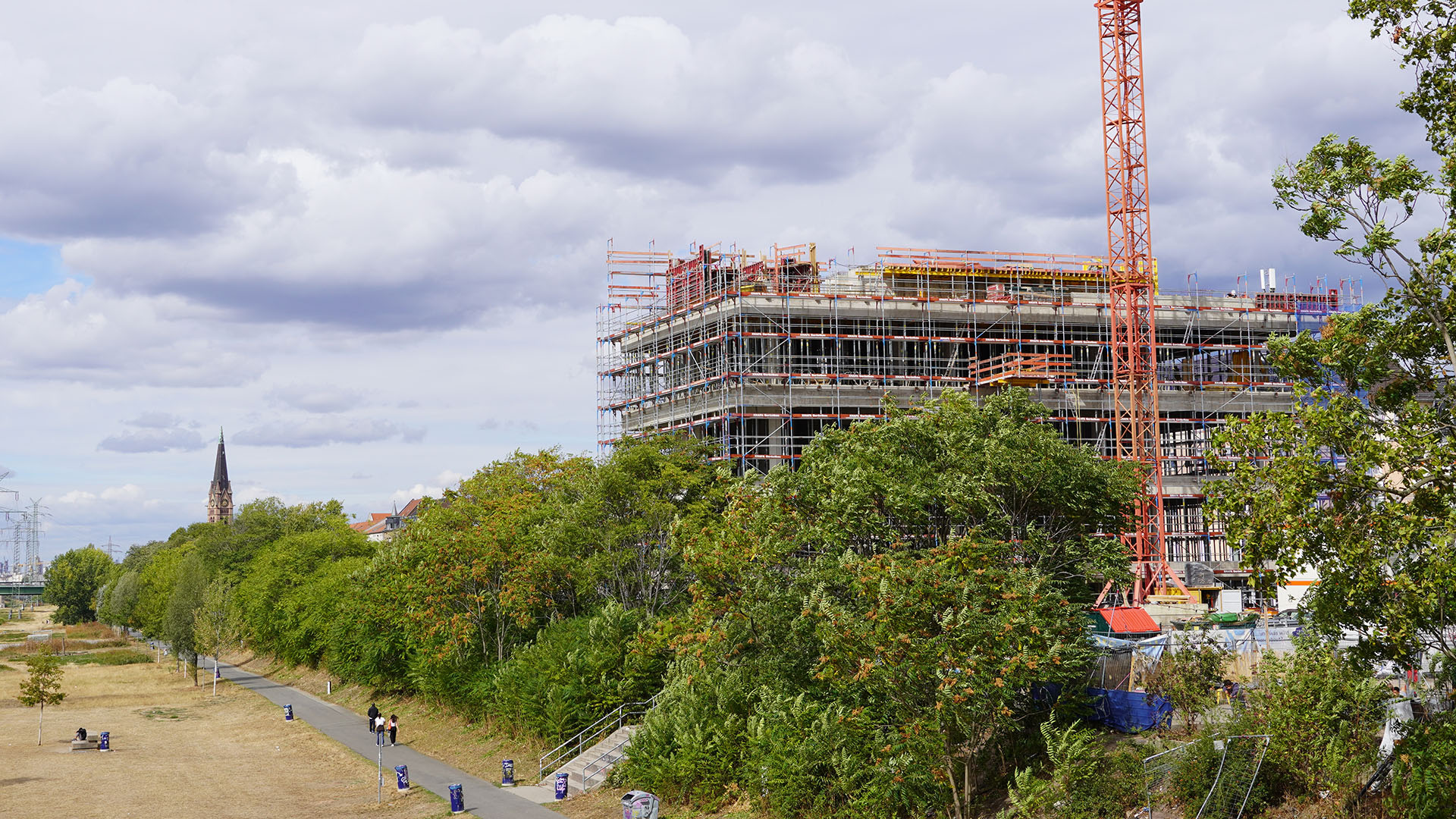 A technologically advanced industrial and research facility under construction, featuring state-of-the-art scaffolding and a crane, situated adjacent to a landscaped path with green trees—reflecting innovative design, sustainable integration, and forward-thinking environments for scientific and educational excellence.