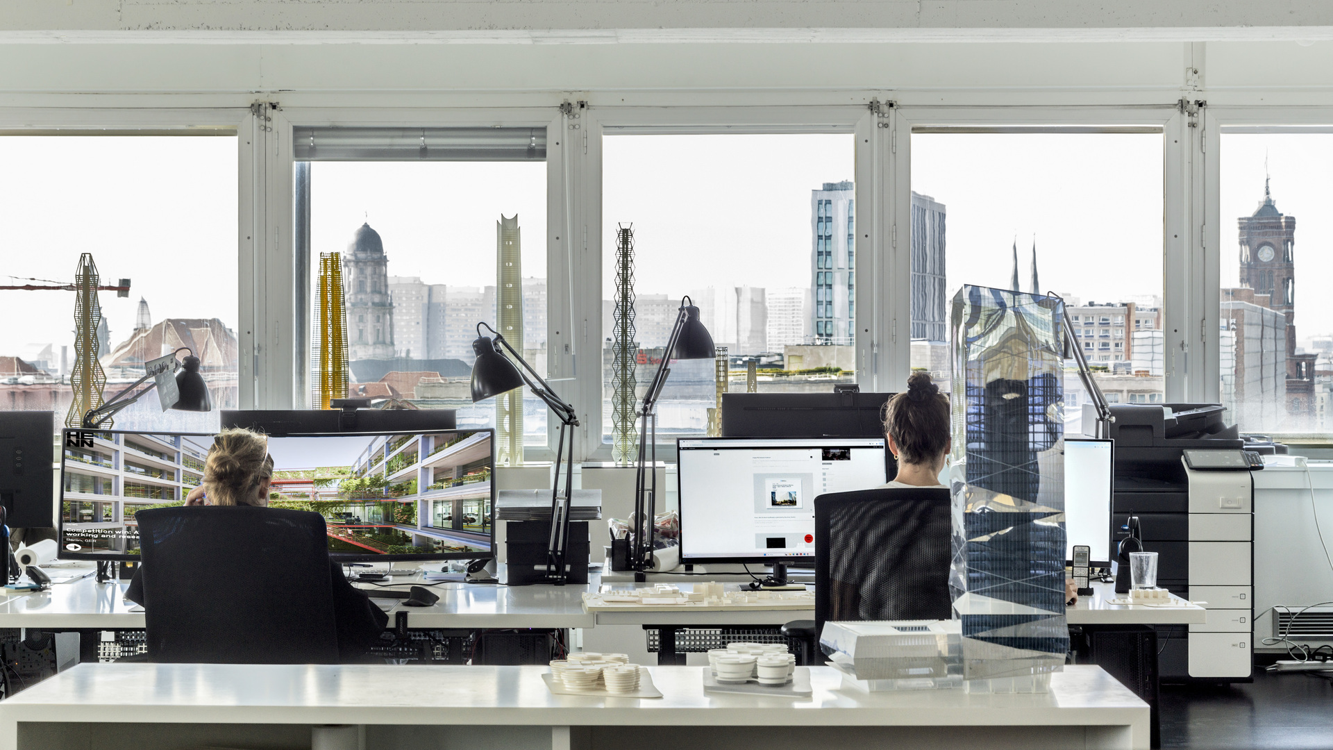 Two professionals work at ergonomic desks with advanced computers in a sleek, high-tech office featuring expansive floor-to-ceiling windows that showcase a vibrant cityscape, exemplifying innovative architectural design focused on sustainability and efficient, technology-driven workspaces ideal for research and industrial applications.