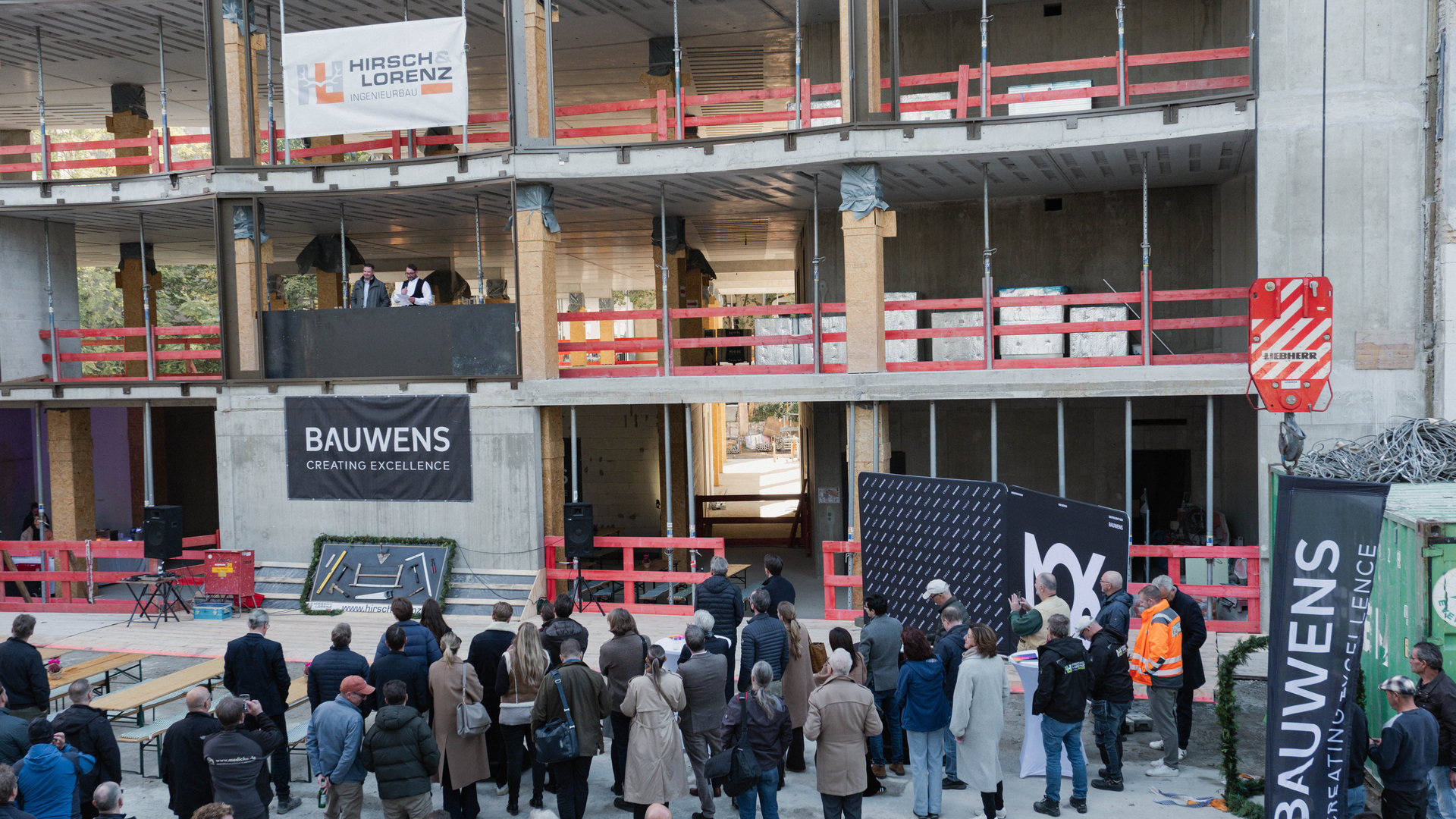 Eine bunte Gruppe von Teilnehmern versammelt sich zu einer Veranstaltung im Freien auf der Baustelle eines mehrstöckigen Gebäudes, um den Fortschritt eines zukunftsweisenden Hightech-Industriegebäudes zu präsentieren.
